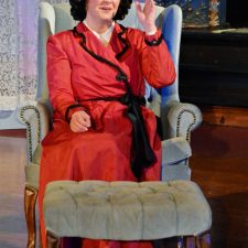 Carrie Kincaid as Ruth sitting in chair in a red dress